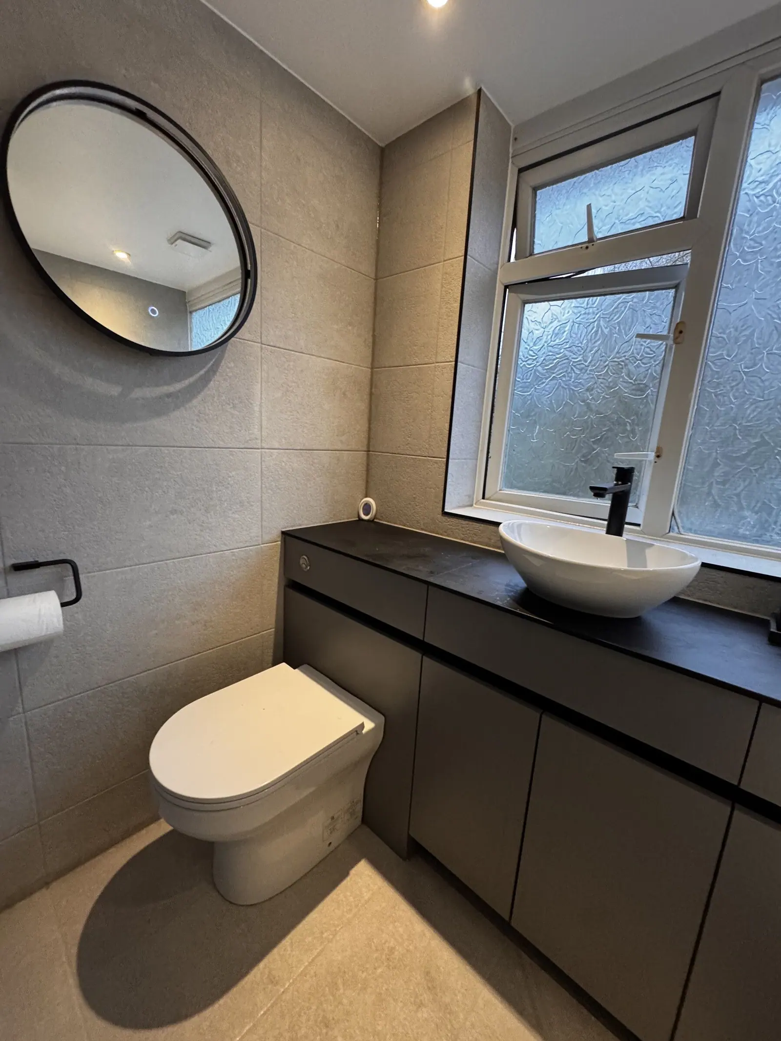 Bathroom with grey tiles, dark handleless vanity, white countertop basin and black-framed round mirror, fitted by Basingstoke Bathrooms